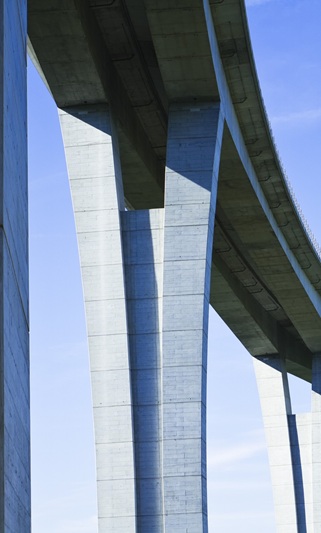 Das Bild zeigt die Unterseite und die massiven Pfeiler einer modernen Brücke aus hochfestem Beton vor blauem Himmel. Die schlanken, elegant geschwungenen Stützen tragen das Brückendeck und verdeutlichen die Leistungsfähigkeit und Gestaltungsfreiheit, die mit hochfestem Beton möglich ist. Hochfester Beton zeichnet sich durch eine besonders hohe Druckfestigkeit (ab ca. 55–60 N/mm²) und ein sehr dichtes, kapillarporenarmes Gefüge aus. Er wird vor allem im Brückenbau, bei Hochhäusern und überall dort eingesetzt, wo Bauteile besonders schlank, tragfähig und langlebig sein müssen